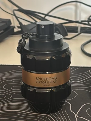 Spicebomb Extreme från Viktor & Rolf - En parfymflaska i svart glas med guldbrun etikett och texten Spicebomb Extreme från Viktor & Rolf. Flaskan har en unik, granatinspirerad form och ett svart spraymunstycke med kedjedetalj. Perfekt för dig som gillar kryddiga och intensiva dofter.