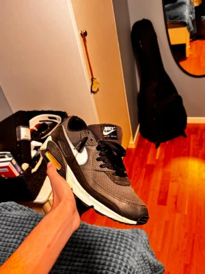 Nike Air Max 90 svart/vit sneaker - Nike Air Max 90 sneakers i svart med vita detaljer och synlig Air Max-dämpning i sulan. Skorna har en klassisk låg siluett, svart mesh och läder på ovandelen samt vit Swoosh-logga på sidorna. Perfekt för dig som gillar streetwear och sportig stil.