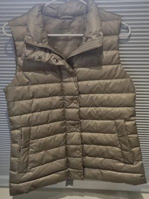 Beige dunväst från GANT - Stilren beige dunväst från GANT med hög krage och tryckknappar framtill. Västen har två fickor med dragkedja och är quiltad i horisontella ränder. Perfekt för lager-på-lager och ger en clean look till din outfit. Köpt för 600 kr och är andvänd 1 gång