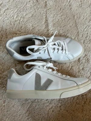 Vita Veja sneakers med gröna detaljer - Snygga vita sneakers från Veja med gröna mockadetaljer på häl och sidor samt klassisk V-logga. Skorna har vita snören och en krämfärgad sula. Tillverkade i läder och mocka för en stilren och trendig look.