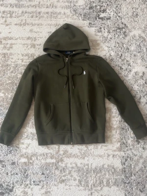 Olivgrön hoodie från Polo Ralph Lauren - Snygg olivgrön hoodie med dragkedja från Polo Ralph Lauren. Klassisk design med huva och dragsko, två fickor framtill och den ikoniska vita loggan broderad på bröstet. Perfekt för en avslappnad och stilren look.