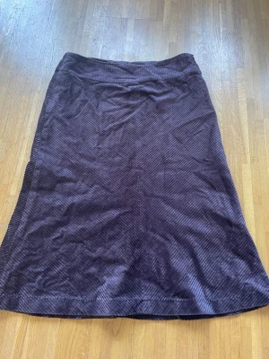 Mörklila manchesterkjol  - Säljer en lång, mörklila manchesterkjol från H&M i storlek 38. Kjolen har en rak och enkel design med lite utsvängt mot slutet. Snedrandigt mönster i tyget. Perfekt för dig som gillar klassiska och stilrena plagg med en twist. Dragkedja på sidan. 