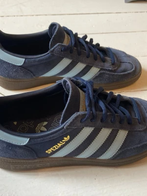Adidas Spezial blå sneakers - Hej säljer ett par snygga Adidas Spezial sneakers i mörkblå mocka med ljusblå ränder och gummisula. Klassisk retrostil med guldfärgad Spezial-logga på sidan. Skorna har låga skaft och matchande blå skosnören. Är knappt använda så är i väldigt bra skick. Hör av er vid frågor eller funderingar 