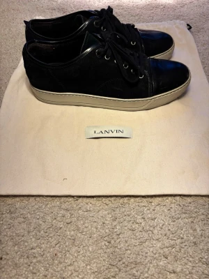 Svarta sneakers från Lanvin - Stilrena svarta sneakers från Lanvin med ovandel i mocka och detaljer i blankt läder. Skorna har svarta skosnören, metallöljetter och en vit sula som ger en snygg kontrast. Perfekta för dig som gillar en clean och lyxig look. Vid några frågor är det bara att höra av er. Nypris kostar dom 5300 kr mitt pris är 2200 kr ( pris kan diskuteras vid snabb affär )