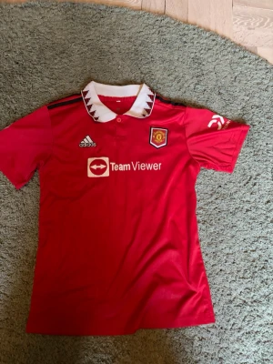 Manchester United matchtröja Antony 21 - Röd Manchester United matchtröja från Adidas med vitt krage och svarta detaljer. Tryck med TeamViewer-logga på bröstet och klubbmärke. På ryggen står det 'ANTONY' och nummer 21. Perfekt för fotbollsfans! Materialet är polyester.