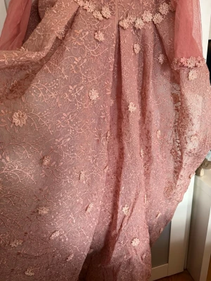 Rosa långklänning med spets och blommor - Romantisk rosa långklänning med transparenta långärmar och vackra spetsdetaljer. Klänningen har broderade blommor och volang vid midjan samt dragkedja i ryggen. Perfekt för speciella tillfällen där du vill sticka ut.