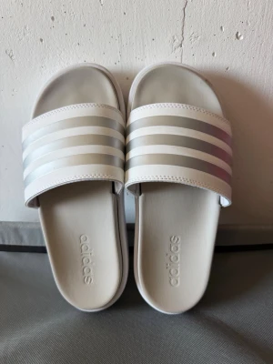 Vita Adidas sandaler med ränder - Snygga vita sandaler från Adidas med klassiska tre silverfärgade ränder över foten. Tillverkade i syntetmaterial med bekväm fotbädd och öppen tå. Perfekta för sommaren och enkel att matcha till olika outfits.