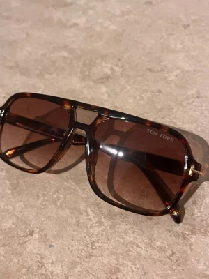 Bruna solglasögon från Tom Ford - Snygga solglasögon från Tom Ford med brunmelerad båge och bruna glas. Modellen har en klassisk fyrkantig form med coola detaljer i guld på sidorna. Perfekt för dig som vill ha en trendig accessoar med exklusiv känsla.
