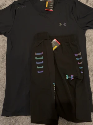  Under Armour - Snyggt svart träningsset från Under Armour med t-shirt och shorts. Båda plaggen har reflekterande, regnbågsskimrande loggor och detaljer. Materialet är lätt och andas, perfekt för träning eller sportaktiviteter. Elastisk midja på shortsen för skön passform.
