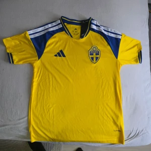 Svensk landslagströja Adidas gul - Gul svensk landslagströja från Adidas med blå detaljer och vita ränder på axlarna. Tröjan har korta ärmar, broderad landslagssköld på bröstet och klassisk sportig design. Perfekt för fotboll eller träning.