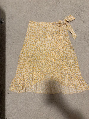 Gul blommig omlottkjol från SHEIN - Säljer en söt gul omlottkjol från SHEIN med vitt och blå blommönster. Kjolen har volangkant och knytband i midjan för en extra fin detalj. Perfekt för varma dagar och ger en fräsch och somrig vibe.