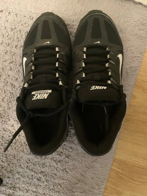 Nike Reax svarta sneakers - Svarta Nike Reax sneakers med vit swoosh-logga på sidan och texten 'TRAINING' på plösen. Skorna har en sportig design med robust sula och svarta snören. Materialet är en mix av syntet och textil, perfekt för en aktiv livsstil.
