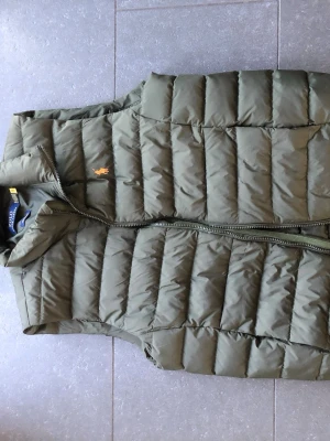 Olivgrön dunväst från Polo Ralph Lauren - Snygg olivgrön dunväst från Polo Ralph Lauren med gul broderad logga på bröstet. Västen har hög krage, dragkedja framtill och quiltad design. Perfekt för lager-på-lager och håller dig varm med stil.