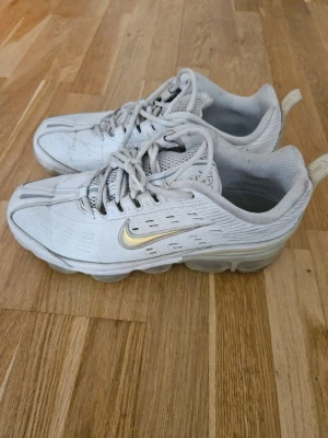 Nike Air Max vita sneakers med guld - Nike Air Max sneakers i vitt med snygga gulddetaljer på sidan och klassisk swoosh. Skorna har en sportig design med mesh och syntetmaterial, samt synlig Air Max-dämpning i sulan. Perfekta för dig som gillar streetwear och vill sticka ut med stilrena detaljer.