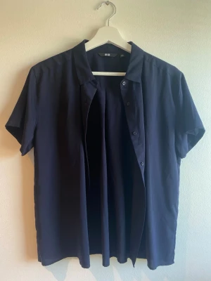 Mörkblå kortärmad skjorta Uniqlo M - Stilren mörkblå skjorta från Uniqlo med korta ärmar och klassisk krage. Skjortan har knappar framtill och är tillverkad i ett mjukt, lätt material som passar perfekt till sommaren. Enkel och clean design utan mönster eller extra detaljer.
