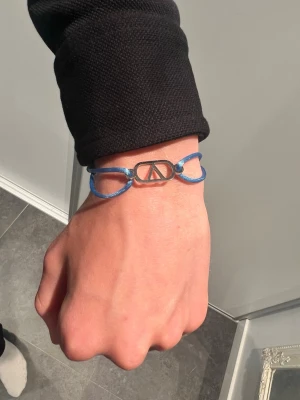 Valentino armband  - Snyggt armband med blått snöre och en silverfärgad metallogo i mitten. Armbandet har en enkel och modern design som passar perfekt till din vardagsstil. Justerbar storlek med knut på snöret.