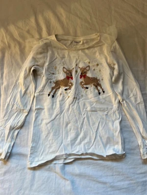 Vit långärmad topp med rådjur Kappahl - Söt vit långärmad topp från Kappahl med tryck av två rådjur med röda rosetter och guldiga stjärnor på bröstet. Perfekt för vintriga dagar och julmys. Tillverkad i mjuk bomull för skön känsla hela dagen. Se sista bild, är en liten fläck på ena armen!