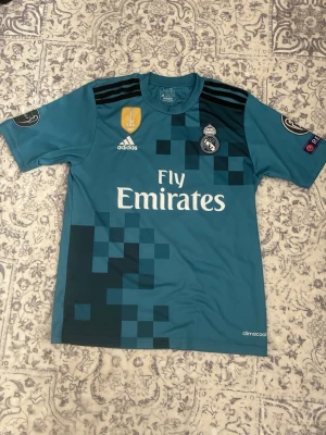 Real Madrid bortatröja Ronaldo #7 - Säljer en blå Real Madrid bortatröja med Ronaldo och nummer 7 på ryggen. Tröjan har pixelmönster, Champions League-patchar på ärmarna och Fly Emirates-tryck på bröstet. Tillverkad i lätt och ventilerande climacool-material. Den är inte äkta men ändå väldigt bra kvalité.
