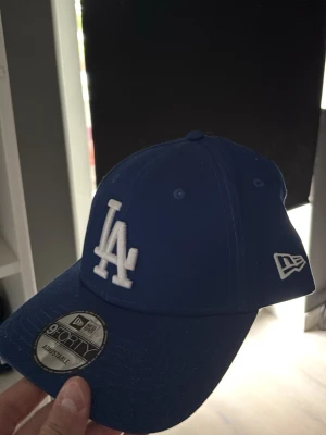 Blå LA-keps från New Era - Snygg blå keps från New Era med LA-logga broderad i vitt framtill och New Era-logga på sidan. Modellen är 9FORTY och har justerbar rem bak. Klassisk böjd skärm och ventilerande hål upptill. Perfekt för dig som gillar streetwear och sportig stil.