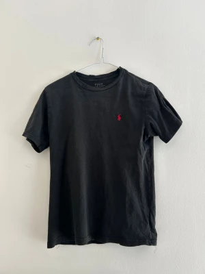 Svart t-shirt från Polo Ralph Lauren - Klassisk svart t-shirt från Polo Ralph Lauren med rund hals och korta ärmar. Liten röd broderad logga på bröstet ger en clean och stilren look. Tillverkad i mjuk bomull som känns skön mot huden. Perfekt basplagg till garderoben. Passar dig som är runt 150-158. Tvättad så det syns men fortfarande fin. Hör av dig vid frågor! 🚀🚀