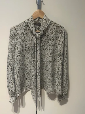 Prickig blus med knytband i halsen - Snygg vit blus med svarta prickar och knytband i halsen. Bluseny har långa ärmar med breda manschetter och knappar framtill. Passar till både jeans och kjol.
