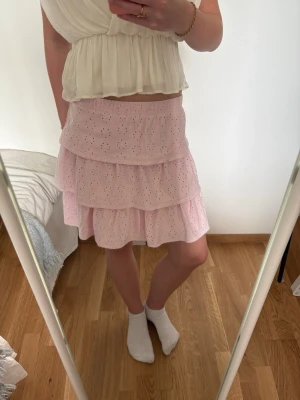 Rosa volangkjol med broderade detaljer - Söt rosa kortkjol med två lager volanger och broderade hål. Kjolen har en elastisk midja och är gjord i ett lätt, luftigt bomullstyg. Perfekt för dig som gillar romantisk och feminin stil.