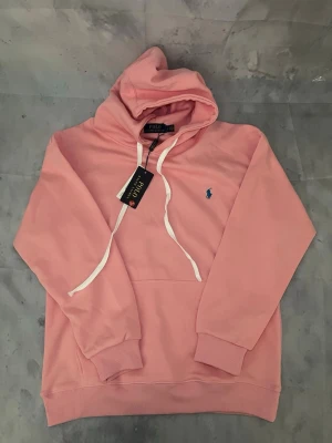 Rosa hoodie från Polo Ralph Lauren - Snygg rosa hoodie från Polo Ralph Lauren med klassisk huva och vita dragsnören. Tröjan har en liten blå logga broderad på bröstet och ribbade muddar vid ärmar och nederkant. Perfekt för en avslappnad och trendig look.