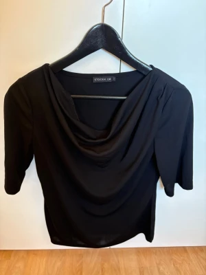 Svart draperad topp från Stockh LM - Stilren svart topp från Stockh LM med draperad halsringning och halvlånga ärmar. Perfekt för dig som gillar en enkel men elegant look. Tillverkad i mjukt material som faller snyggt över kroppen. Nypris 600