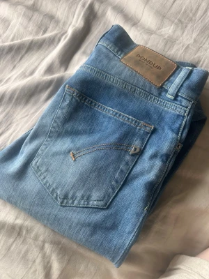 Ljusblå jeans från Dondup - Säljer ett par ljusblå jeans från Dondup med klassisk femficksdesign och läderpatch bak i midjan. Jeansen har raka ben och är tillverkade i mjukt denimtyg. Perfekta för en avslappnad och trendig look.