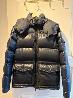 Svart pufferjacka från Moncler - Snygg svart pufferjacka från Moncler med hög krage och två stora fickor framtill med lock. Jackan har glansig finish, dragkedja och logga på dragkedjan. Perfekt för kalla vinterdagar och riktigt varm tack vare den tjocka vadderingen.