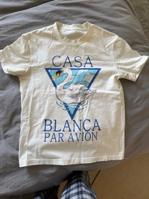 Vit t-shirt Casablanca  - Vit t-shirt med tryck framtill som visar en svan och texten 'Casa Blanca Par Avion' i blått. T-shirten har klassisk passform och rund hals. Perfekt för dig som gillar unika prints och stilrena färger.