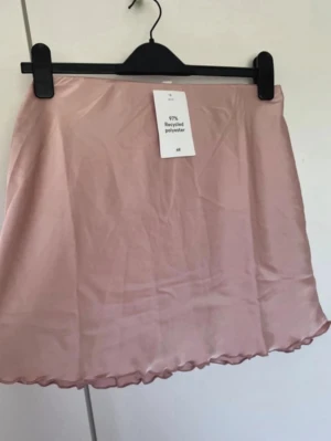 Ljusrosa kjol från H&M i satin - Ljusrosa kjol från H&M i satinliknande polyester. Kjolen har en enkel, A-linjeformad design med lätt volangkant nedtill och hög midja. Materialet är 97% återvunnen polyester och ger ett mjukt, glansigt intryck. Perfekt för dig som gillar stilrena och trendiga plagg.