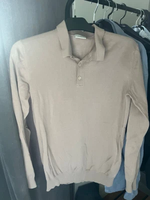 Beige långärmad pikétröja John Henric - Snygg beige långärmad pikétröja från John Henric. Tröjan har klassisk krage och två knappar framtill. Tillverkad i tunt, mjukt material som ger en clean och stilren look. Perfekt för dig som gillar minimalistisk stil. Använd ett fåtal gånger och riktigt fin beige färg! 