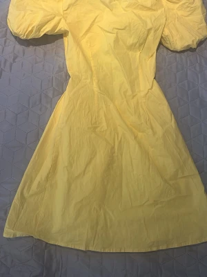 Gul kortklänning med puffärmar - Snygg gul kortklänning med puffärmar och öppen rygg. Klänningen har en omlottdetalj baktill och är tillverkad i ett lätt bomullsliknande material. Perfekt för soliga dagar och ger en fräsch och trendig look.