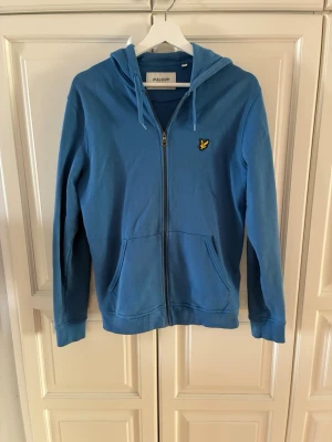 Blå zip-hoodie från Lyle & Scott - Snygg blå hoodie med dragkedja från Lyle & Scott. Klassisk modell med huva och fickor framtill samt den ikoniska gula loggan på bröstet. Tillverkad i mjukt bomullsmaterial, perfekt för en avslappnad stil.