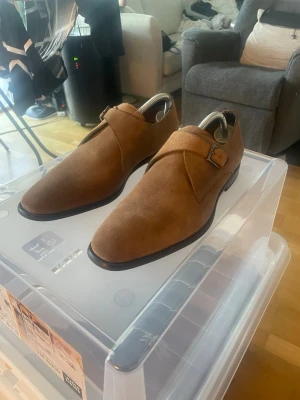 Bruna mocka loafers från Digel - Sprillans nya, snygga bruna loafers i mocka från Digel med klassisk monk strap-detalj och stilren design. Skorna har en smal siluett, låg klack och spänne i metall på sidan. Perfekta för dig som vill ha en clean och modern look. 