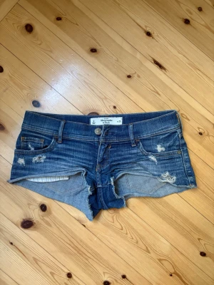 Blå jeansshorts Abercrombie & Fitch - Säljer ett par blå denimshorts från Abercrombie & Fitch med slitna detaljer och råa kanter. Klassisk femficksmodell med låg midja och knappgylf. Perfekta för varma dagar och en avslappnad look. Super söta och perfekta till sommaren💕🌺