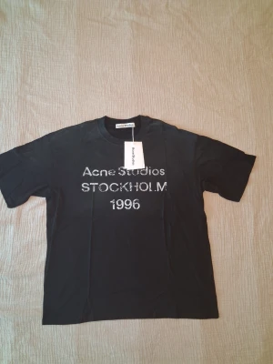 Svart Acne Studios t-shirt med tryck - Cool svart t-shirt från Acne Studios med texten 'Acne Studios STOCKHOLM 1996' i vitt framtill. Klassisk passform med rund hals och korta ärmar. Perfekt för en stilren och trendig look.