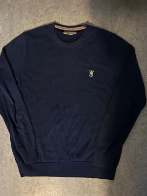 Mörkblå tröja från Burberry - Mörkblå rundhalsad tröja från Burberry med broderad logga på bröstet. Tröjan har ribbade muddar vid ärmslut och nederkant samt en klassisk passform. Halsringningen har en snygg detalj med ränder i flera färger. Perfekt för en stilren look.