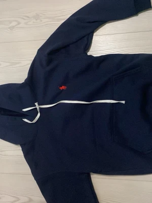 Mörkblå hoodie från Polo Ralph Lauren - Mörkblå hoodie från Polo Ralph Lauren med vit dragsko och klassisk röd logga broderad på bröstet. Hoodien har känguruficka framtill och är tillverkad i mjukt material som känns skönt mot huden. Perfekt för en avslappnad och stilren look.