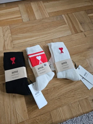 3-pack Ami strumpor med hjärtdetalj - Kan köpas individuellt också. Säljer ett 3-pack ribbade strumpor från Ami. Ett par är svarta, ett par vita med röda ränder och ett par helvita. Alla har Ami:s ikoniska röda hjärta med A broderat på skaftet. Perfekta för att ge din outfit en cool touch.