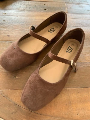 Bruna  ballerinaskor - Bruna ballerinaskor i mockaimitation från 157 med klassisk Mary Jane-rem och fyrkantig tå. Skorna har en justerbar rem med liten metallspänne och en enkel, stilren design som passar till många olika outfits.