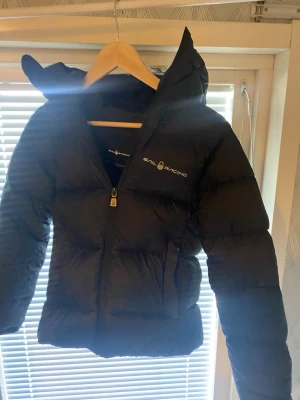 Svart dunjacka från Sail Racing - Säljer en svart dunjacka från Sail Racing med huva och dragkedja framtill. Jackan har logga på bröstet och en rund vit detalj på huvan. Puffig modell som håller dig varm under kalla dagar. Perfekt för dig som vill ha en stilren och funktionell vinterjacka. Jag köpte den här jackan i vintras med den kom inte så mycket till användning, därför är den i väldigt bra skicka. Det är i modellen ”W down hood cloud”