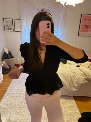 Svart omlottblus med volanger - Säljer en svart omlottblus med volangdetaljer och markerad midja. Blusen har v-ringning och puffiga långärmar i ett tunt, lätt transparent material. Perfekt för dig som vill ha en trendig och feminin look.