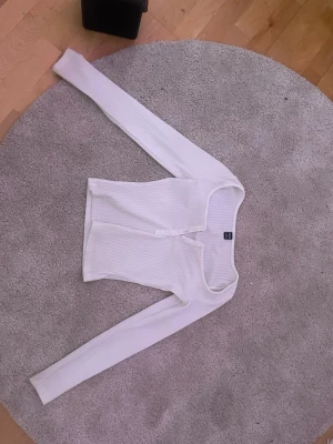 Vit ribbad långärmad topp från H&M - Säljer en vit långärmad med ribbad struktur och knappdetaljer framtill. Toppen har en snygg, lite djupare rundad halsringning och är gjord i ett mjukt, stretchigt material som sitter skönt på kroppen.