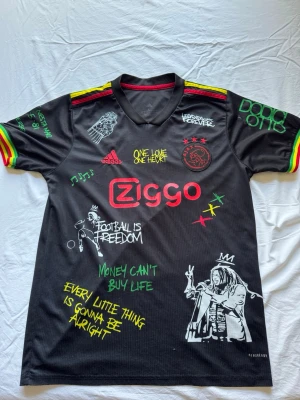 Ajax x Bob Marley fotbollströja - Snygg svart Ajax fotbollströja från Adidas med Bob Marley-tema. Tröjan har färgglada detaljer i rött, gult och grönt på ärmarna, tryckta citat och illustrationer. Kan gå ner vid snabb affär!