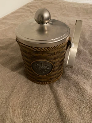 Isbehållare i flätad korg med tång - Snygg isbehållare i brun flätad korg med silverfärgat lock och handtag. Medföljer en matchande is-tång i metall. Behållaren har en dekorativ metallplatta med druvmotiv på framsidan. Perfekt till festen eller hemmabaren.