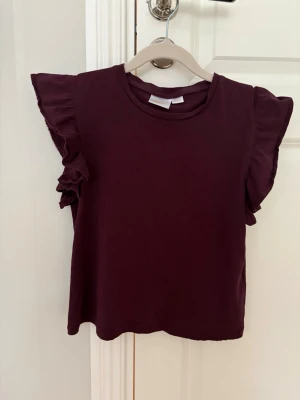 Vinröd topp med volangärmar Gina Tricot - Supersöt vinröd topp från Gina Tricot med volangdetaljer på ärmarna. Rund halsringning och rak passform. Perfekt för dig som vill ha en enkel men ändå trendig look.