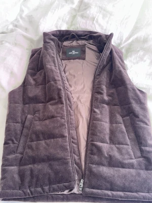 Brun manchesterväst John Henric - Snygg brun dunväst från John Henric i mjuk manchester. Västen har dragkedja framtill, två sidofickor och quiltad design. Perfekt för lager på lager och ger en stilren look till din outfit.