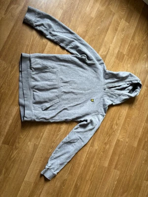 Lyle & Scott - Säljer en grå hoodie från Lyle & Scott med deras klassiska broderade bi-logga i gult och svart på bröstet. Hoodien har huva med dragsko och en stor magficka framtill. Tillverkad i mjuk bomullsblandning och har ribbade muddar vid ärmar och nederkant. Enda att anmärka på är lite nött vid armar se bild.